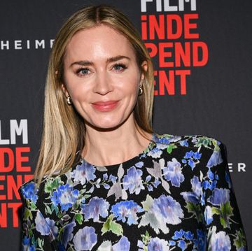 emily blunt smiles at the camera, she wears a blue and silver floral patterned dress and stands in front of a black background with writing