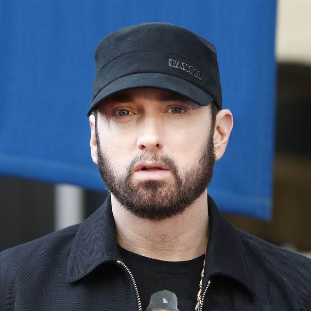 eminem looks at the camera with a straight face, he wears a black cap and jacket with a black graphic t shirt and golden chain necklace