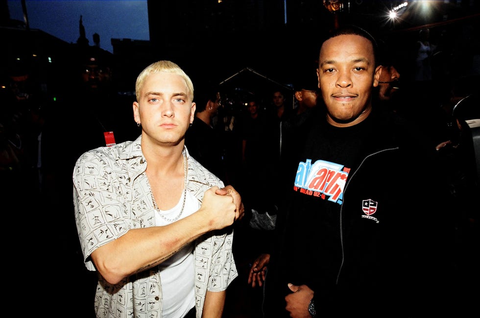 eminem and dr dre clasp hands while standing next to each other and looking at the camera, eminem wears an unbutton patterned collared shirt over a white tank top, dre wears a black zip up jacket over a black graphic tshirt