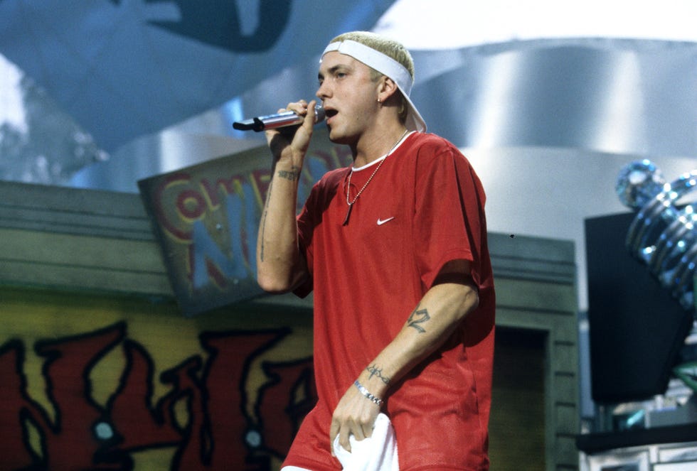 eminem raps into a wireless microphone he holds in one hand, he wears a backward white visor, red tshirt, dog tag, and silver bracelet