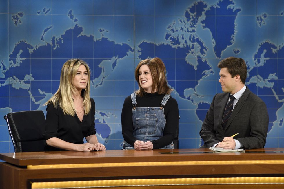 jennifer aniston, vanessa bayer, and colin jost sit at a wooden desk in front of a two tone blue world map