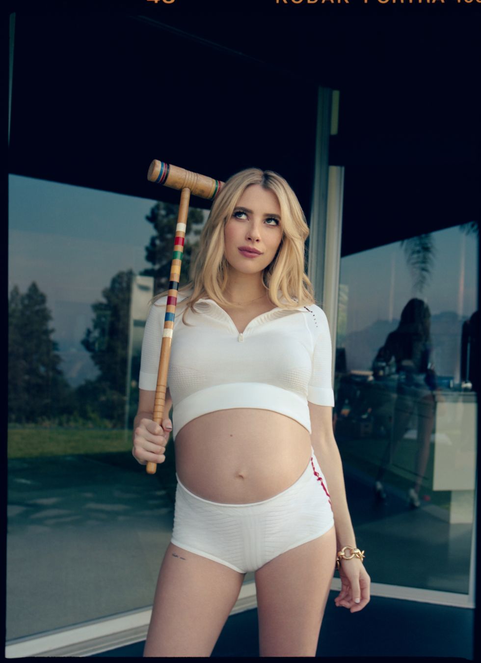 emma roberts, pregnant, wearing white crop top and shorts, holding uo croquet mallet next to head, standing in front of windows of home