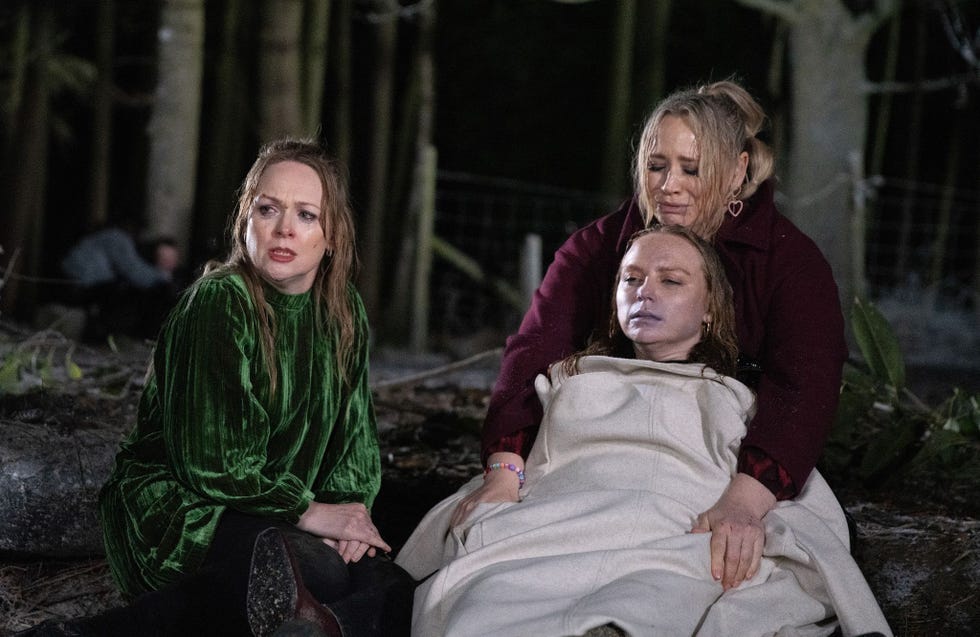 three individuals seated in a dark forest after a crash, one looks very ill being covered by a coat