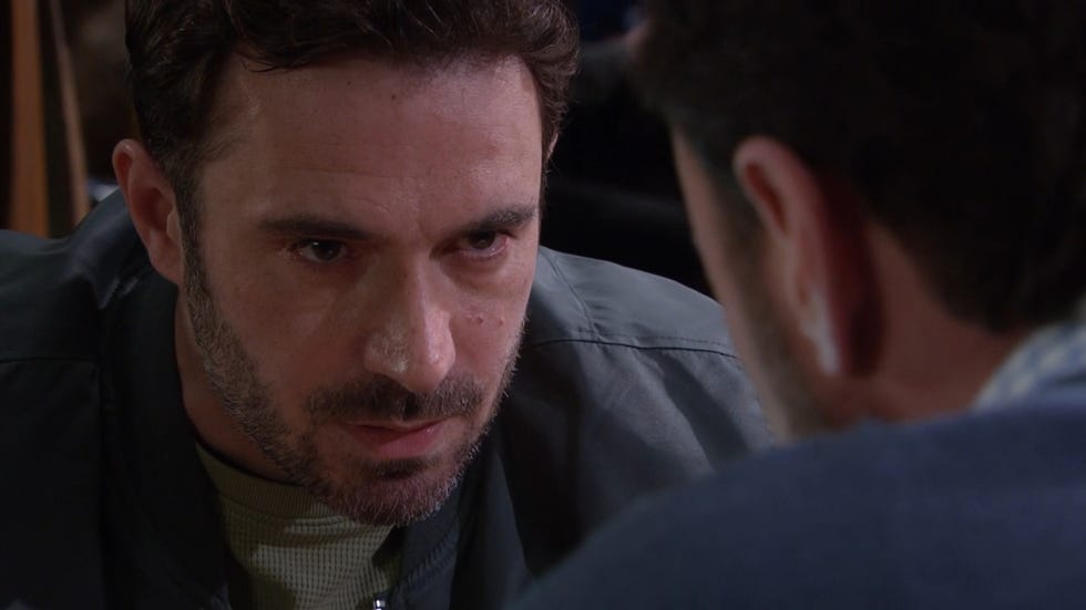 emmerdale, a close up of john sugden in a dark blue jacket and green jumper, with an angry look on his a face staring at dr liam cavanagh, who is out of focus