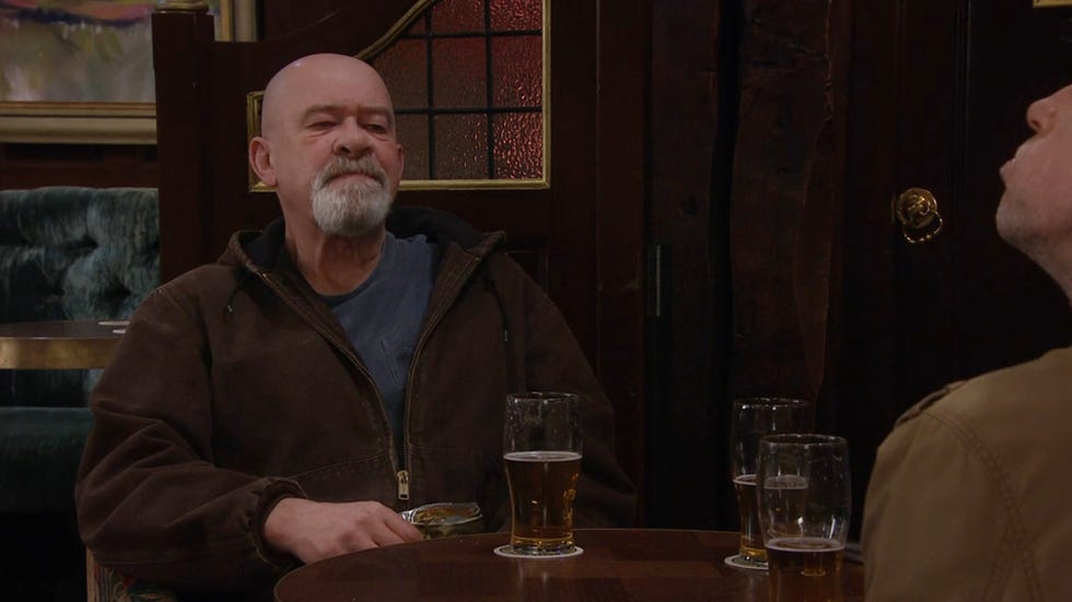 emmerdale, bear wolf sitting in the pub with a pink, wearing a dark brown zip up jacket over a blue tshirt, looking unimpressed
