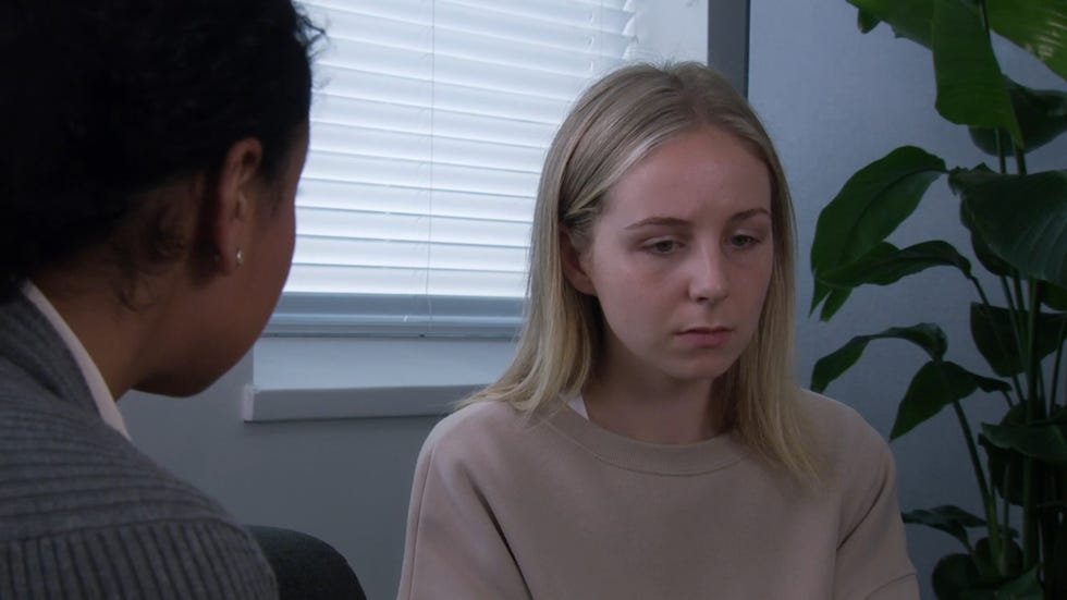 emmerdale, belle king in a very gray room with a plant behind her