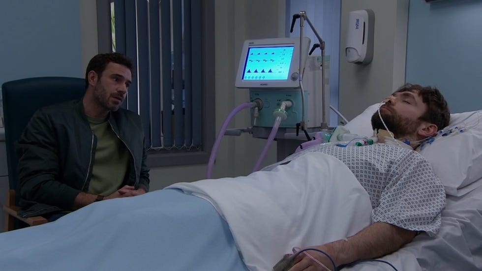 emmerdale, john sugden in a hospital wearing a dark blue jacket and green jumper, sat down talking to a man with a brown hair in a coma lying in bed