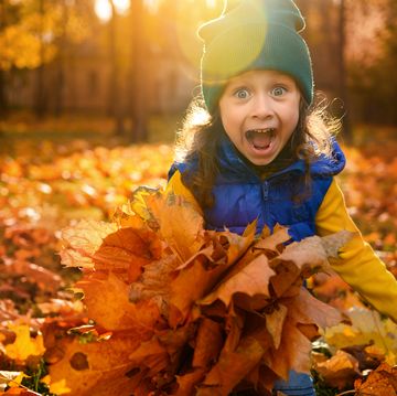 emotional lifestyle portrait of adorable cheerful baby girl in colorful clothes playing with dry fallen autumn maple leaves in golden park at sunset with beautiful sunbeams falling through trees
