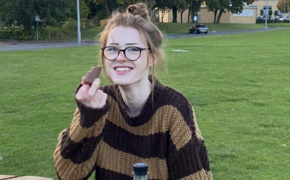 brianna ghey, a teenager sat holding a chocolate bar and wearing a knitted striped jumper she looks happy