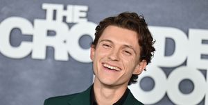 tom holland, wearing a green suit, smiles and looks off camera, standing in front of a logo for the television series the crowded room