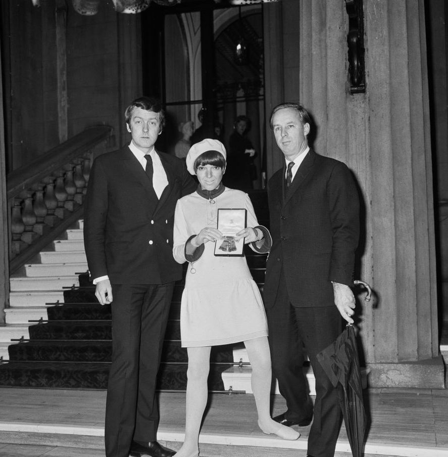 alexander plunket greene, mary quant, and archie mcnair stand inside buckingham palace, both men wear suits, and mary is wearing a mini dress, beret, tights and flats while holding a medal inside a box, mcnair is holding a large closed umbrella, behind them is a large staircase and stone column