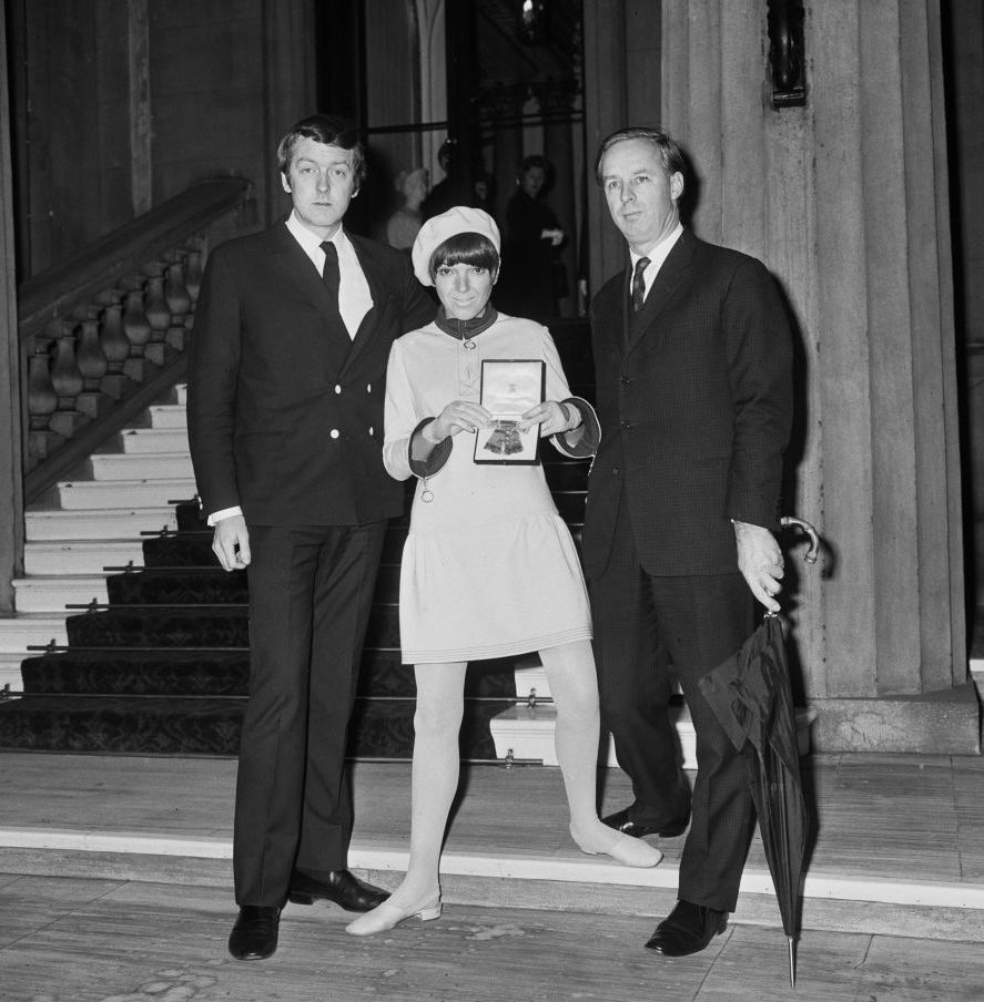 alexander plunket greene, mary quant, and archie mcnair stand inside buckingham palace, both men wear suits, and mary is wearing a mini dress, beret, tights and flats while holding a medal inside a box, mcnair is holding a large closed umbrella, behind them is a large staircase and stone column
