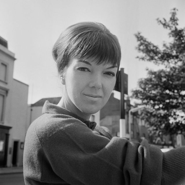 mary quant in a black and white photo from september 1960, she is looking at the camera with a neutral expression and wears a sweater, her hair is in a updo with her bangs reaching just to her eyebrows, she is standing outside on a nondescript london street
