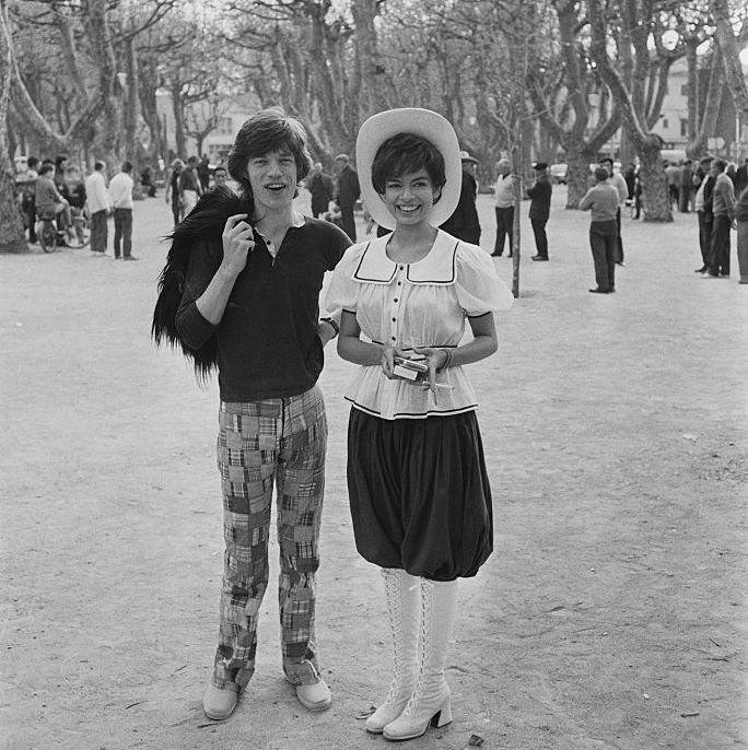 mick jagger and bianca perez moreno de macias smile while standing next to each other outside in front of a field of trees and other people, he wears plaid pants and a dark shirt and holds a shag jacket over one shoulder, she wears a hat, light colored blouse with a collar, dark pants and knee high lace up boots