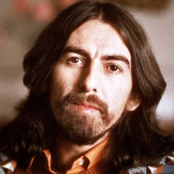 george harrison looks at the camera with a solemn expression, he has shoulder length brown hard, large eyebrows, a full beard and mustache, at the bottom of the image you can just see his orange collared shirt and patterned sweater that is black, white, and gray with pops of orange and soft blue