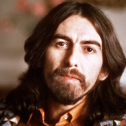 george harrison looks at the camera with a solemn expression, he has shoulder length brown hard, large eyebrows, a full beard and mustache, at the bottom of the image you can just see his orange collared shirt and patterned sweater that is black, white, and gray with pops of orange and soft blue