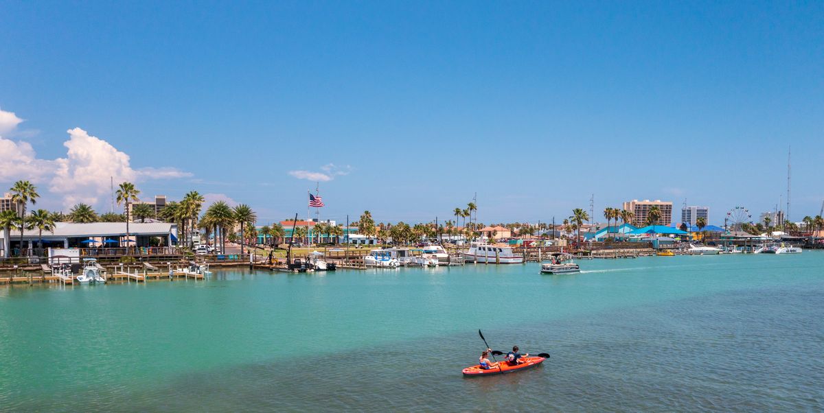 person kayaking on the ocean next to the shore covered with palm trees, boats, businesses and more