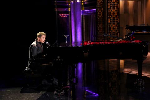 jeremy renner plays piano and sings into a microphone on a television set, there is an empty martini glass and rose petals on top of the piano