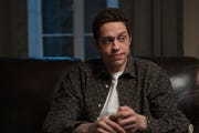 pete davidson wearing a black and white buttoned shirt and a white t shirt, sitting on a couch