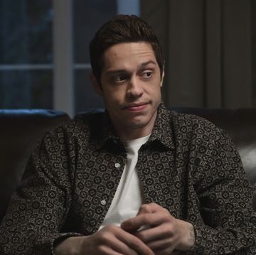 pete davidson wearing a black and white buttoned shirt and a white t shirt, sitting on a couch