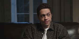 pete davidson wearing a black and white buttoned shirt and a white t shirt, sitting on a couch