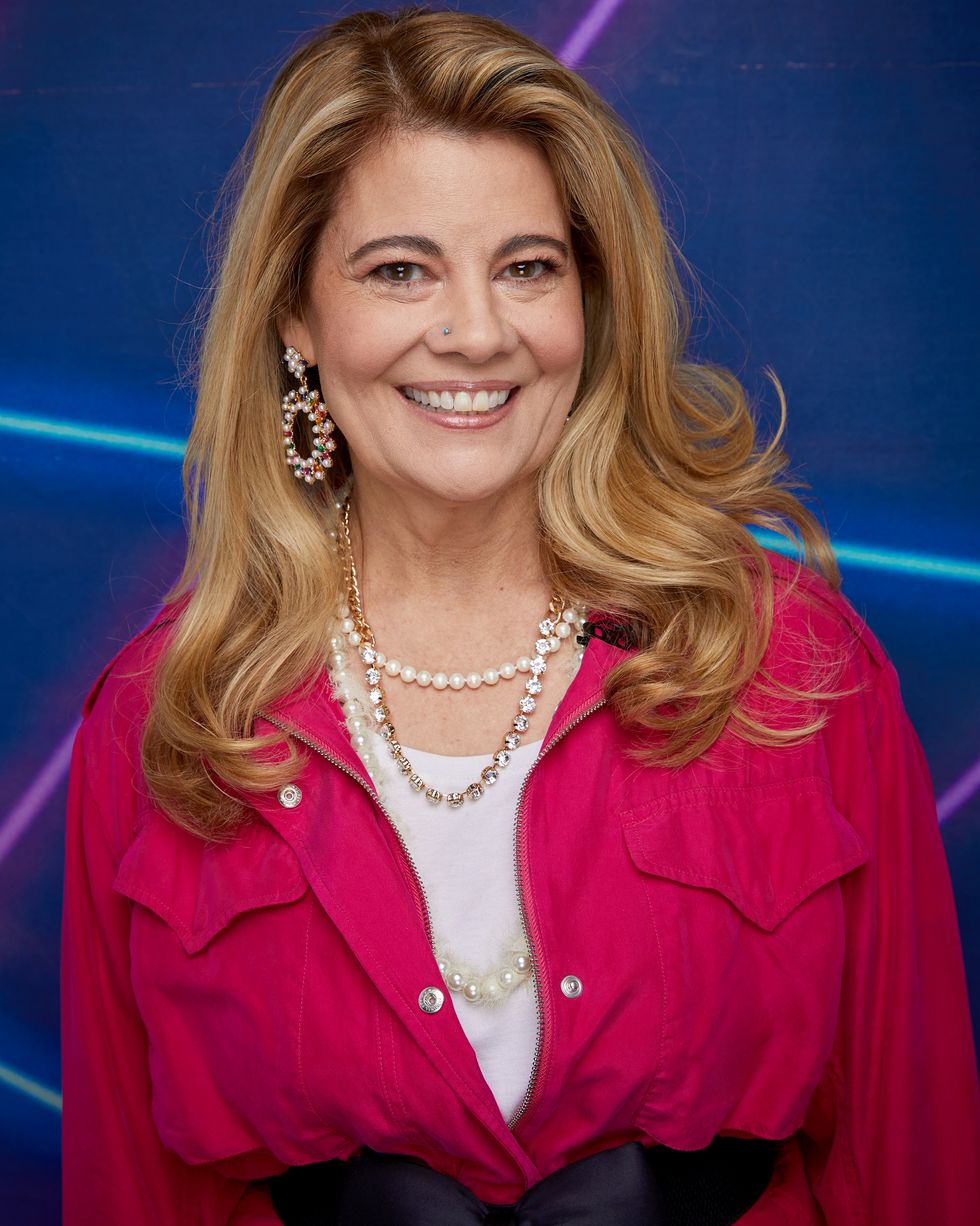 lisa whelchel smiles at the camera, she wears a pink jacket with a bow around the waist and a white shirt as well as several necklaces and large pearl earrings