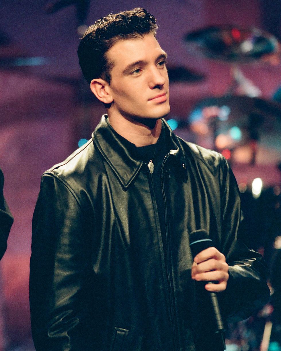 jc chasez holds a microphone at his chest and looks to the right, he wears a black jacket over a black shirt