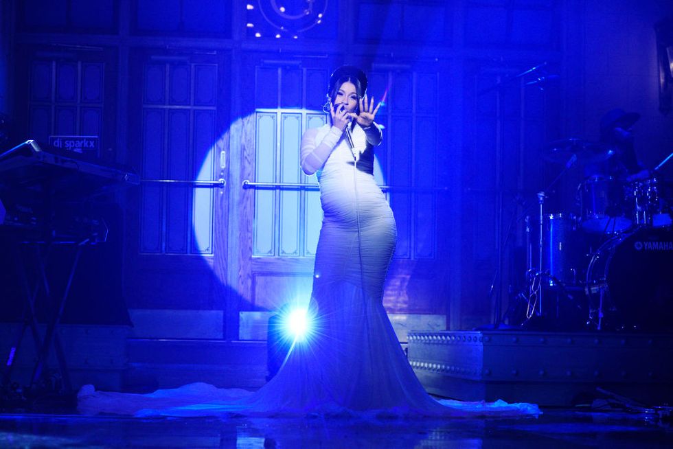 cardi b singing into a microphone on a stage while wearing a white dress that shows her pregnant belly