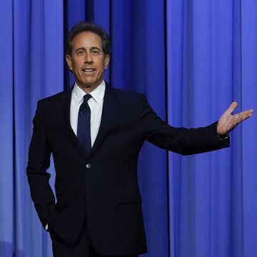 jerry seinfeld stands in front of a blue curtain and extends one arm out, he wears a dark suit with a white collared shirt and tie