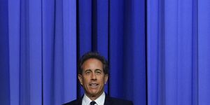 jerry seinfeld stands in front of a blue curtain and extends one arm out, he wears a dark suit with a white collared shirt and tie