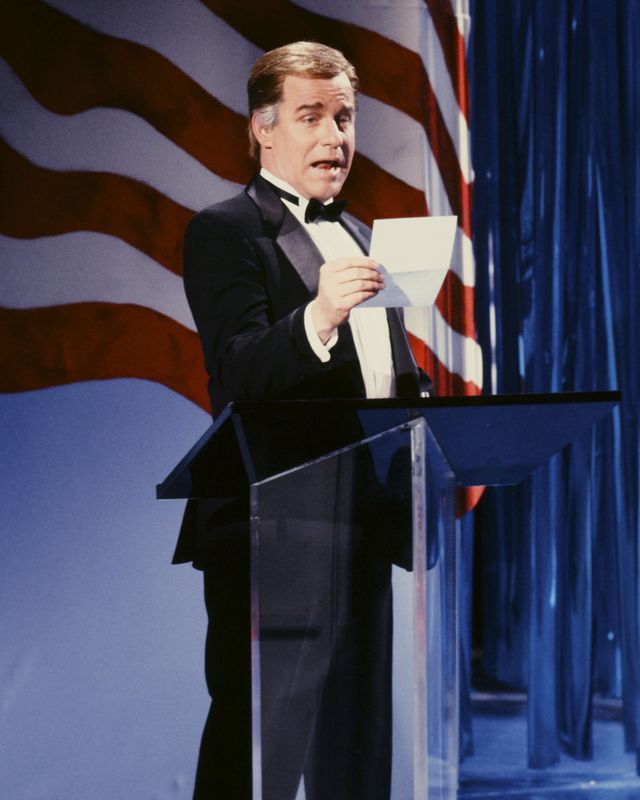 phil hartman wears a black tuxedo and stands at a clear podium, he speaks and holds a piece of paper, behind him is part of a large american flag