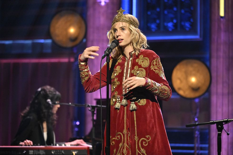 maya hawk sings into a microphone on a stand as she holds her hands in front of her and looks past the camera, she wears a gold fabric crown with a red velvet coat with gold embellishments