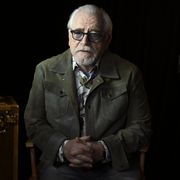 brian cox wearing an olive coat and sunglasses, standing next to a stage light and trunk, with his hands crossed