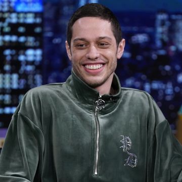 pete davidson smiles and looks down past the camera, he wears a velvet sage green quarter zip jacket, behind him is a blurry tv set of a city skyline