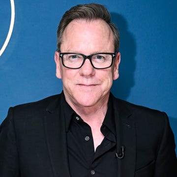 Kiefer Sutherland in December 2023 kiefer sutherland smiles at the camera, he wears black glasses, a black suit jacket and a black collared button up shirt