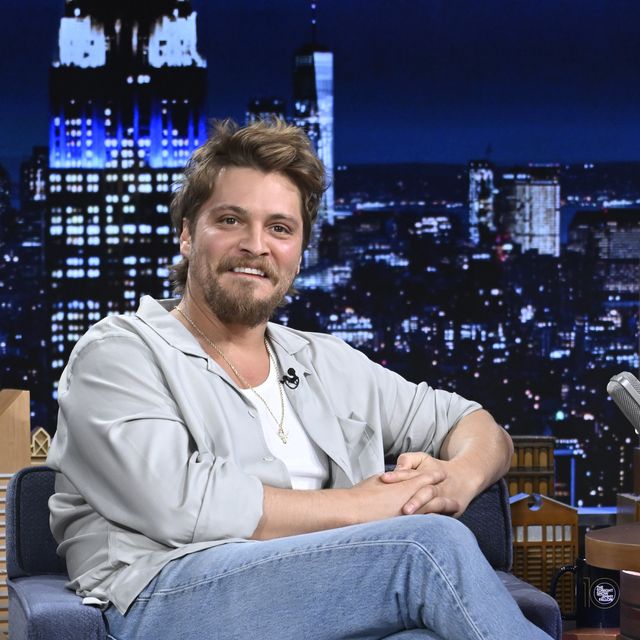 luke grimes smiles as he sits on a tv set in an armchair
