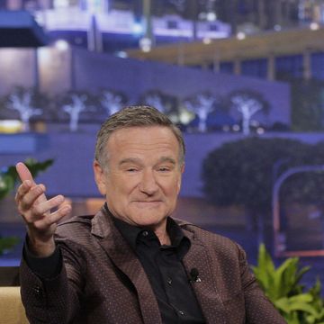 robin williams extends his right arm out toward the camera, he has a slight smile on his face, he wears a brown polka dot suit with a black collared shirt, he is sitting on a mustard yellow couch