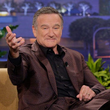 Robin Williams in August 2013 robin williams extends his right arm out toward the camera, he has a slight smile on his face, he wears a brown polka dot suit with a black collared shirt, he is sitting on a mustard yellow couch