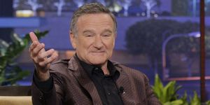 robin williams extends his right arm out toward the camera, he has a slight smile on his face, he wears a brown polka dot suit with a black collared shirt, he is sitting on a mustard yellow couch
