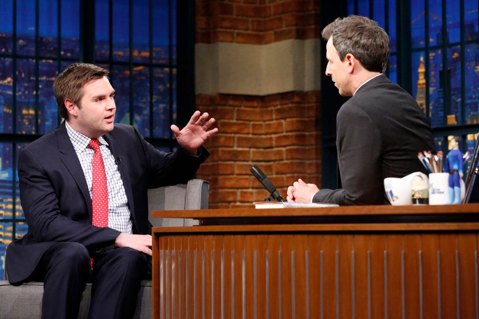 jd vance sits in an armchair and looks to the right at seth meyers who sits behind a desk and looks at vance, both men wear suits