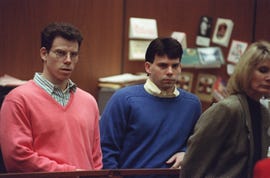 erik menendez and lyle menendez inside a wood paneled room, erik wears glassed and a salmon colored sweater over a green and white striped collared shirt, lyle wears a blue sweater over a yellow and white striped collared shirt