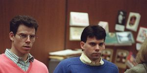 erik menendez and lyle menendez inside a wood paneled room, erik wears glassed and a salmon colored sweater over a green and white striped collared shirt, lyle wears a blue sweater over a yellow and white striped collared shirt
