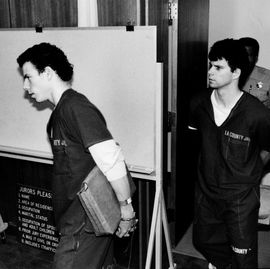 two men walk into a room with their hands handcuffed behind their backs as a police office follows them, a white board stands in the background