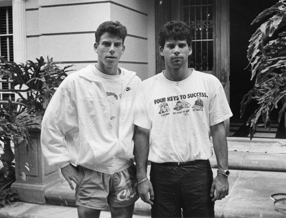 erik and lyle menendez stand outside a home and stare at the camera, the front door is open, erik wears a jacket over a tshirt with shorts, lyle wears a tshirt with pants