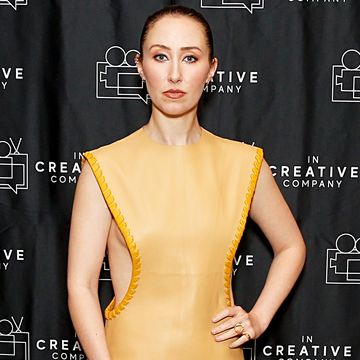 erin doherty, standing with left hand on hip looking at the camera, wearing a yellow dress, dark hair pulled back in a bun