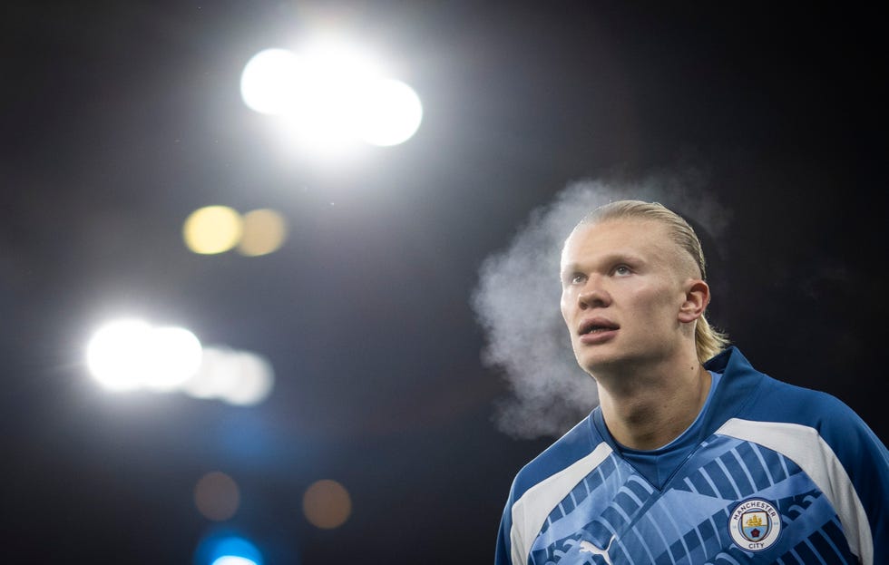 erling haaland stands in a blue manchester city jersey at night with lights in the background