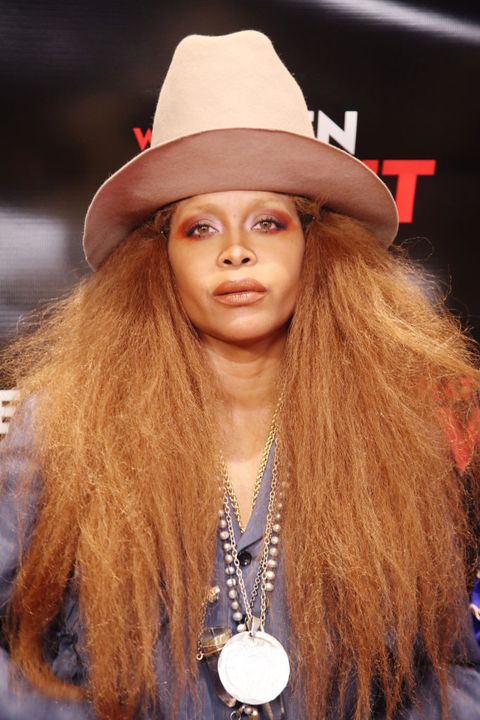 erykah badu poses on the red carpet, wearing a loose button up top with many long necklaces she's wearing a tall, brimmed hat with her hair down, and red eye shadow