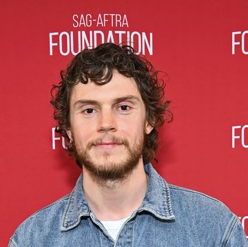 evan peters with a beard wearing a denim shirt and smiling in front of a red background