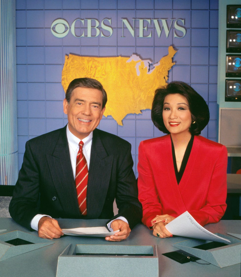 dan rather and connie chung sit at a news desk and smiles, each holds papers in front of them and wears suits