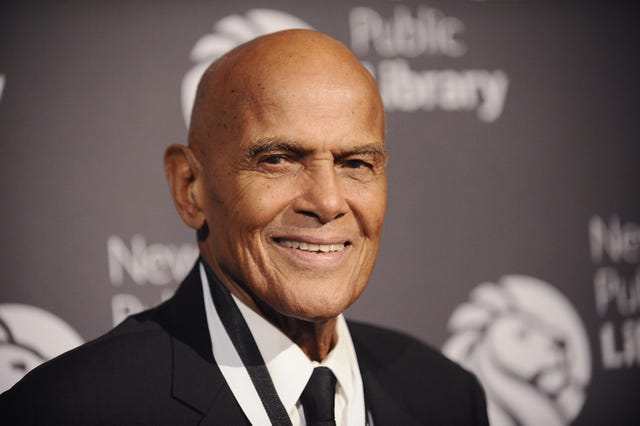 harry belafonte looks toward the camera and smiles, he is wearing a black suit with a white shirt and black tie